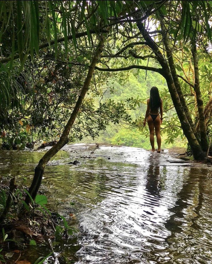 a woman standing in the middle of a river surrounded by trees and foliage, with her back turned to the camera