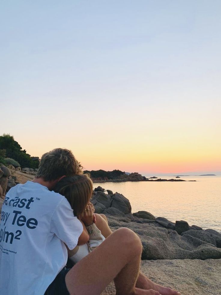 two people sitting on the beach watching the sun set over the water and rocks in front of them