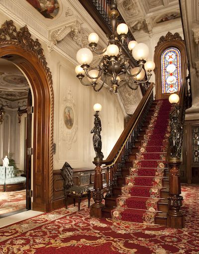 an ornate staircase with red carpet and chandelier in the center, leading up to another room