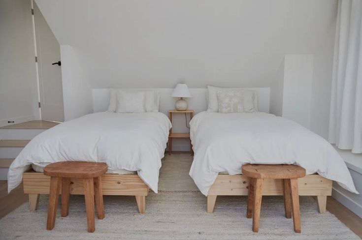 two beds with white linens and wooden foot stools in a room that is painted white