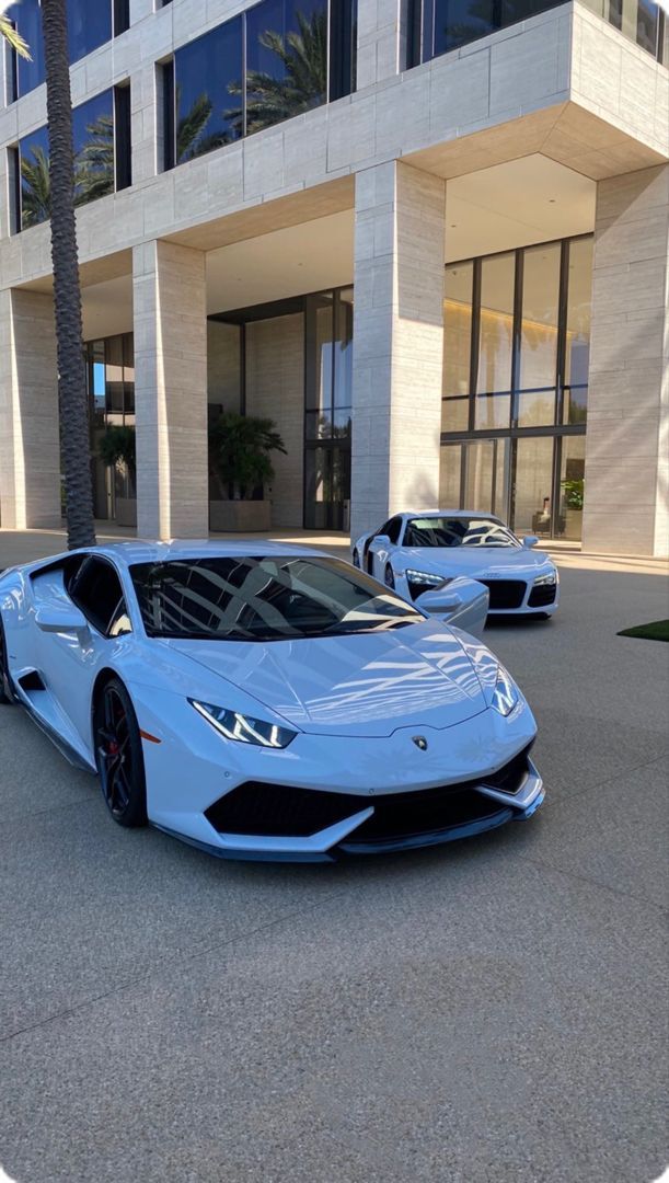 two blue sports cars are parked in front of a large white building with palm trees