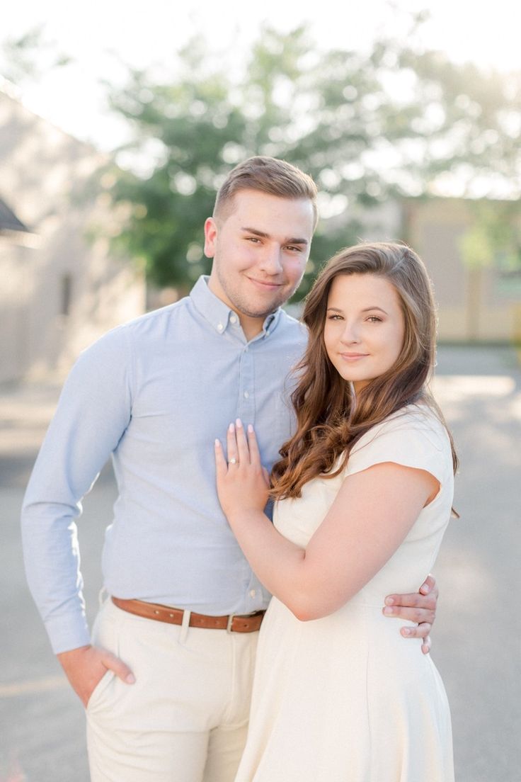 a man and woman standing next to each other in front of a tree with their arms around each other