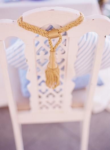 a white chair with a gold tassel on it's seat and some pillows in the background