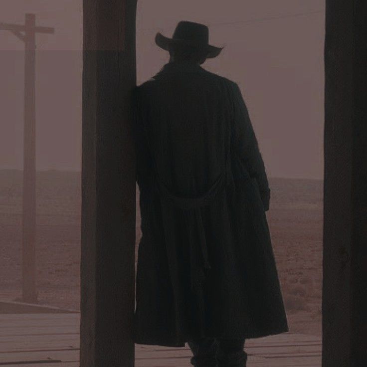 a man in a long black coat and hat looking out the window at an empty field