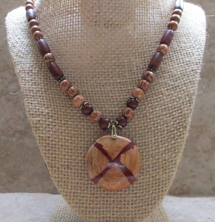 What an eye-catching wood pendant necklace! This one-of-a-kind design will wow you.  The light-colored wood is lamented and splashed with a wine-colored design. The necklace is accented with wine-colored seed beads and palmwood beads. *Necklace length: 21 inches *Pendant size: 1.5 inches in diameter   *Accent beads: Seed and wood *Dark-brown cotton cord *Antique brass pinch bail *Antique brass clasp Mahogany Wooden Beads Jewelry, Mahogany Colored Wooden Bead Jewelry, Gift Brown Wooden Beaded Necklaces, Brown Wooden Beaded Necklace Gift, Wooden Brown Beaded Necklace Gift, Necklace With Beads, Light Colored Wood, Wood Jewelry, Wood Pendant