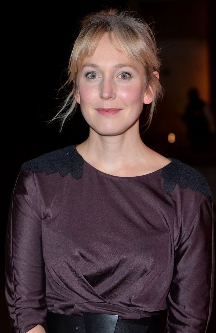 a woman with blonde hair wearing a brown dress and black belted skirt, smiling at the camera