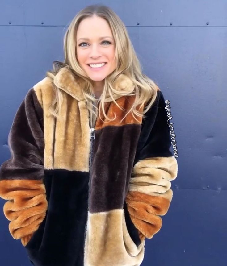 a woman wearing a brown and black jacket standing in front of a blue wall with her hands on her hips