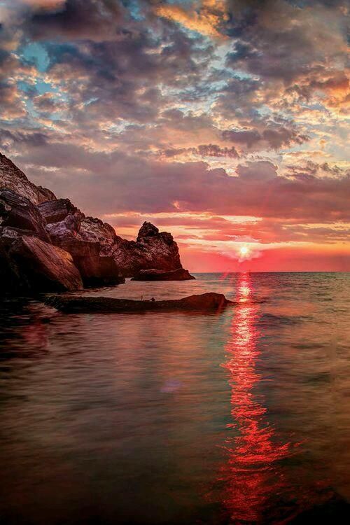 the sun is setting over the ocean with rocks in the foreground and clouds in the background