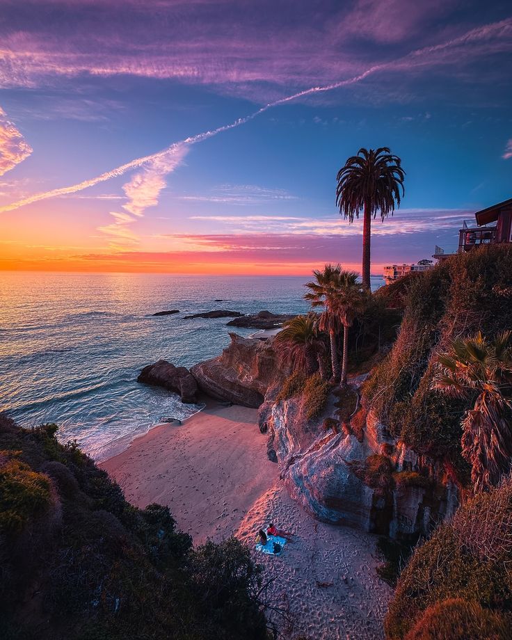 the sun is setting over the ocean with palm trees on the shore and people laying in the sand