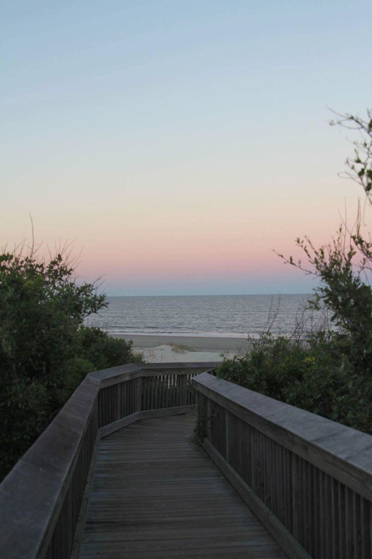 a wooden walkway leading to the beach at sunset with trees on either side and water in the distance