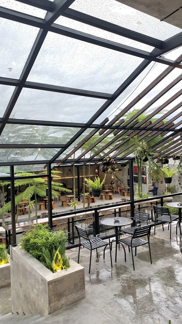 an outdoor dining area with tables and chairs under a glass roof, surrounded by potted plants