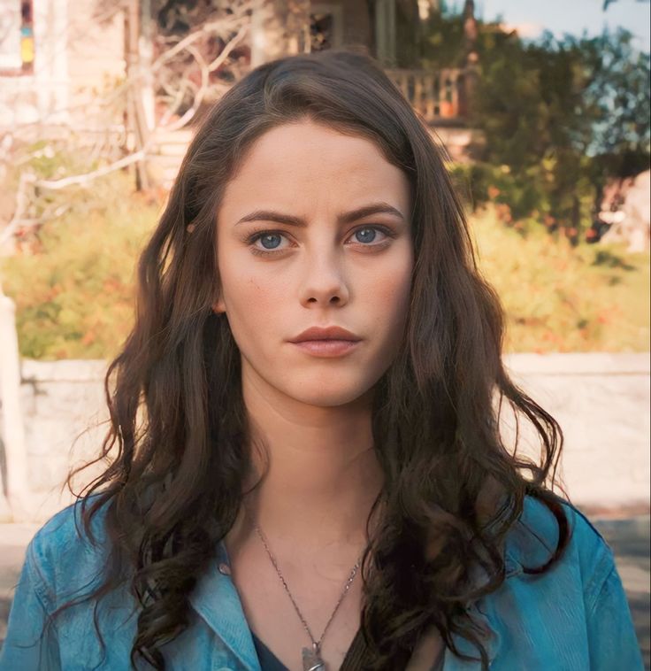 a woman with long brown hair and blue eyes looks at the camera while standing in front of a house