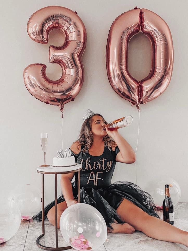 a woman sitting on the floor drinking from a bottle next to balloons and a cake