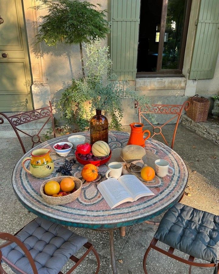 an outdoor table with books and fruit on it in front of a door, next to two chairs