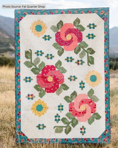 a white quilt with flowers on it in the middle of a grassy field and mountains