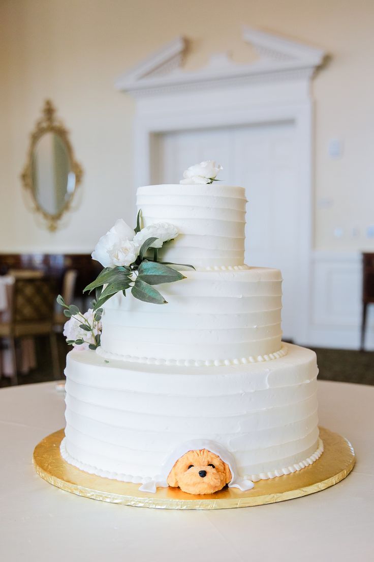 a three tiered white cake with flowers on the side and a teddy bear decoration