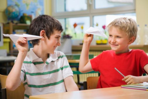 two young boys sitting at a table with paper airplanes in their hands and one boy holding up a piece of paper