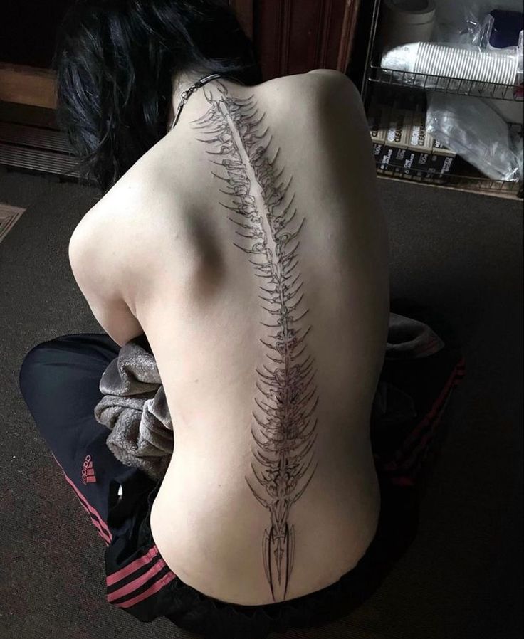 a woman sitting on the floor with her back turned to the camera, showing an intricate fern tattoo