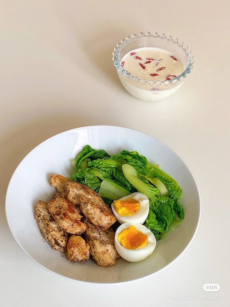 a white plate topped with meat and vegetables next to a bowl filled with eggs, broccoli