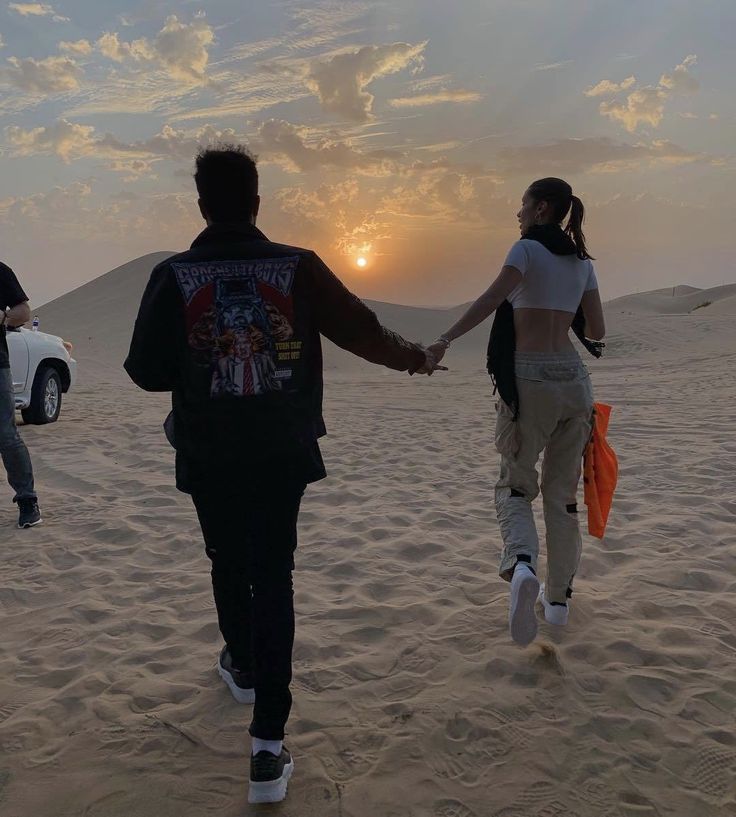 two people holding hands while walking in the sand at sunset with cars parked on the beach behind them