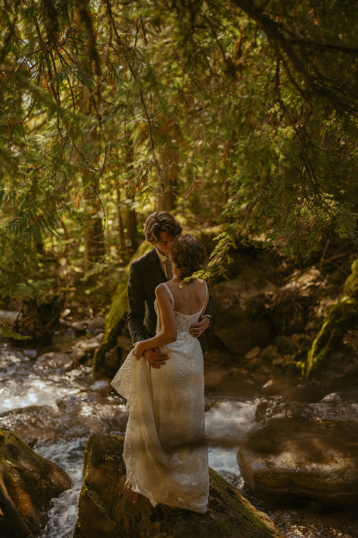 a bride and groom are standing on rocks in the woods near a stream, kissing