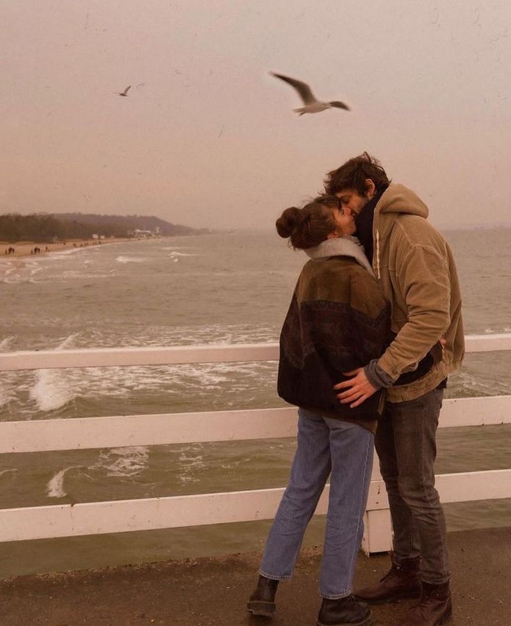 two people standing next to each other near the ocean and seagulls flying overhead