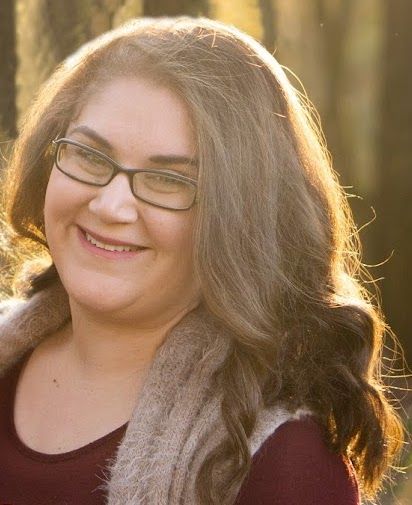 a woman wearing glasses and a scarf smiles at the camera with trees in the background