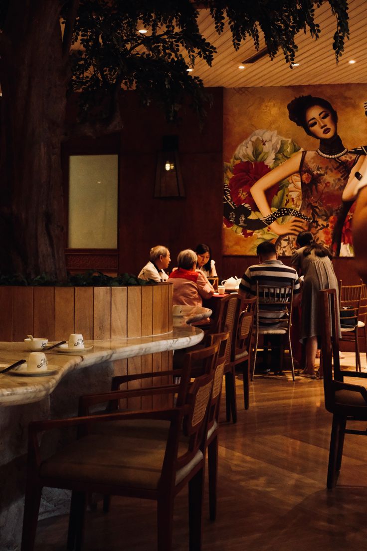 people sitting at tables in a restaurant with paintings on the wall behind them and wooden chairs