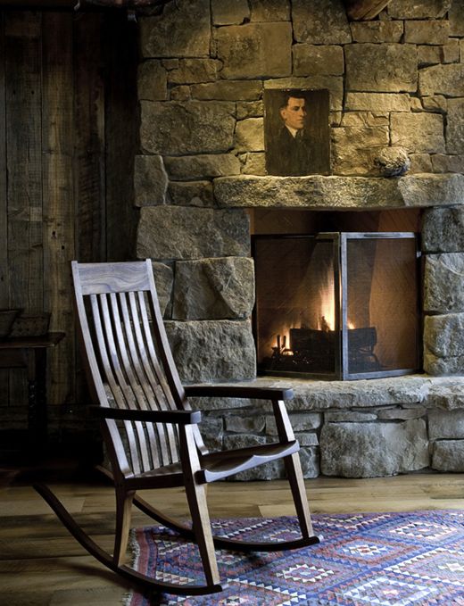 a rocking chair sitting in front of a stone fireplace with a portrait on the mantle