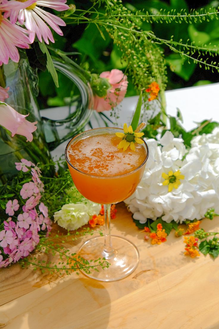 an orange drink sitting on top of a wooden table next to white and pink flowers