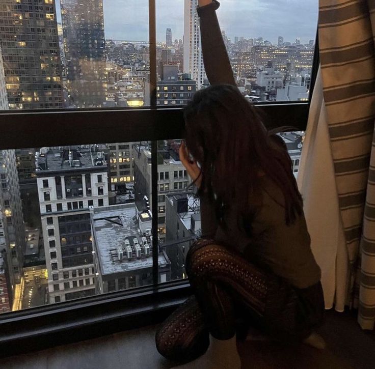 a woman sitting in front of a window looking out at the city lights and skyscrapers