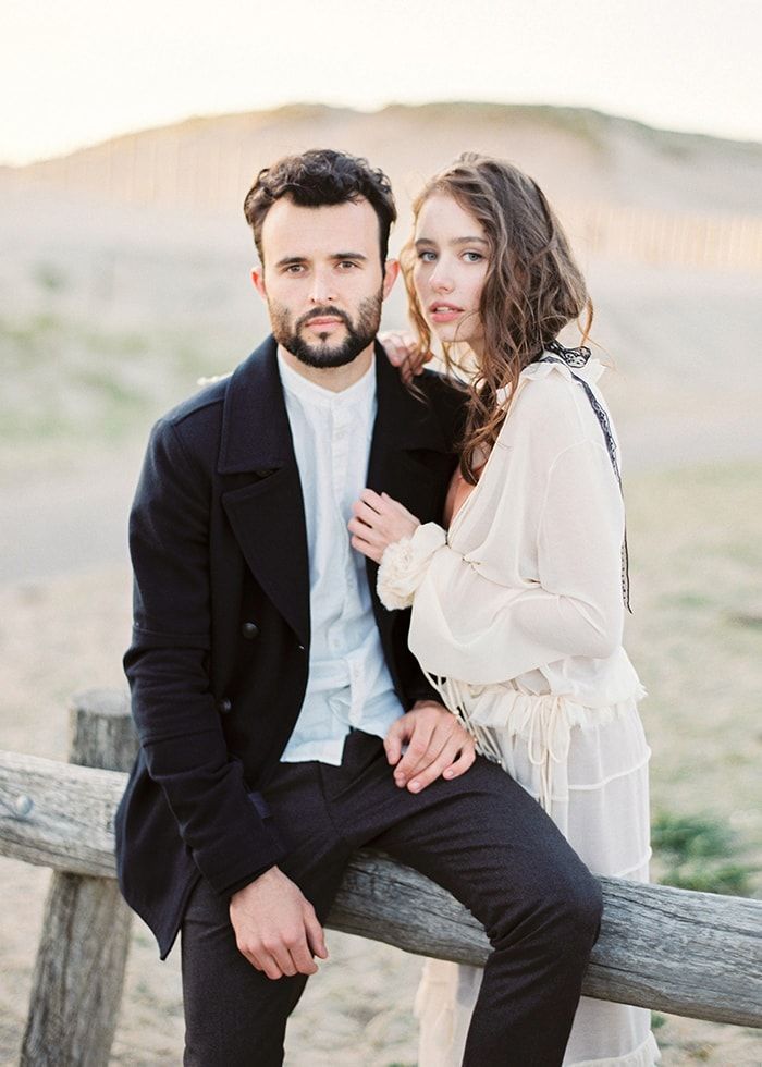 a man and woman sitting on a wooden bench together, posing for a photo in front of a fence