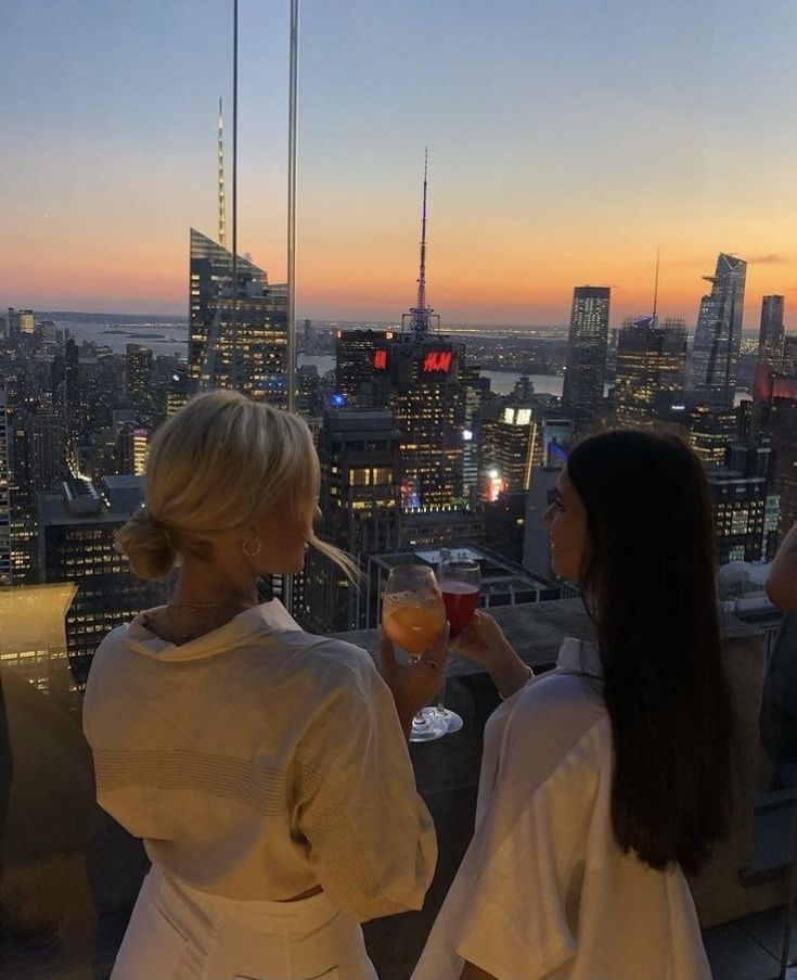 two women standing on top of a building with drinks in their hands and looking at the city