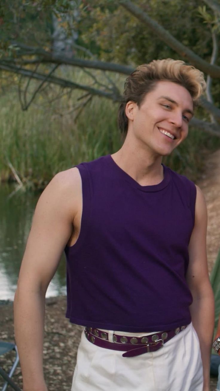 a young man standing in front of a body of water wearing a purple shirt and white shorts