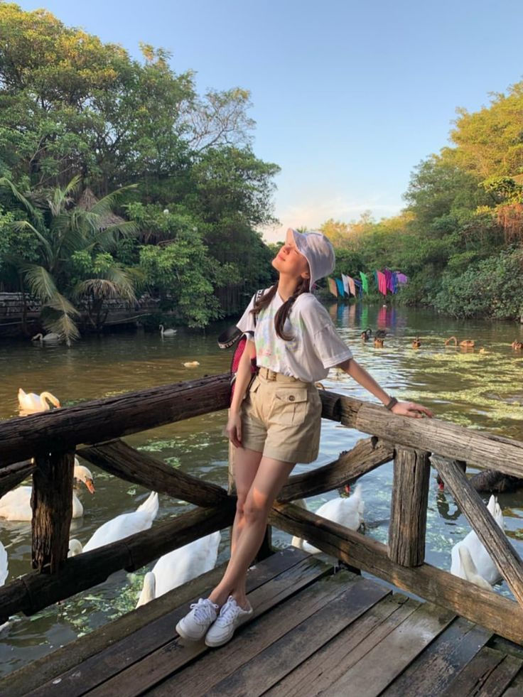 a woman standing on a wooden bridge over water with swans swimming in the river behind her