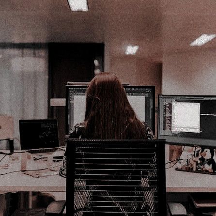 a woman with long red hair sitting at a desk in front of two computer monitors