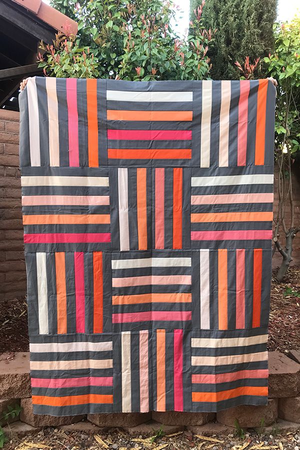 an orange, pink and grey quilt hanging on a brick wall next to some bushes