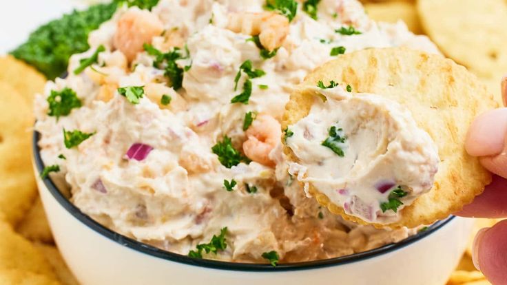 a hand holding a cracker over a white bowl filled with shrimp salad and tortilla chips