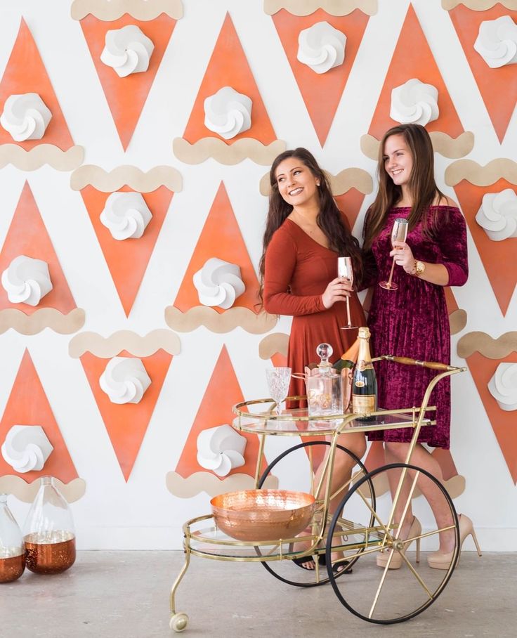 two women standing in front of a wall with orange and white paper decorations on it