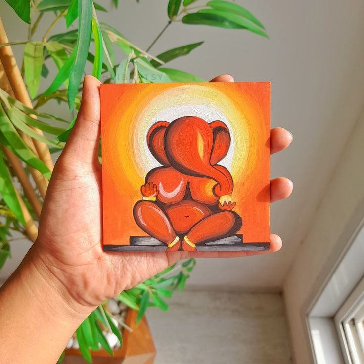 a person holding up a small painting in front of a potted plant