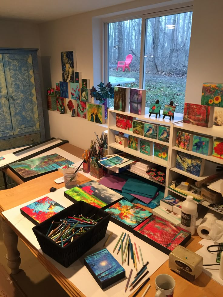 an art studio with many paintings and drawings on the desk, along with two large windows