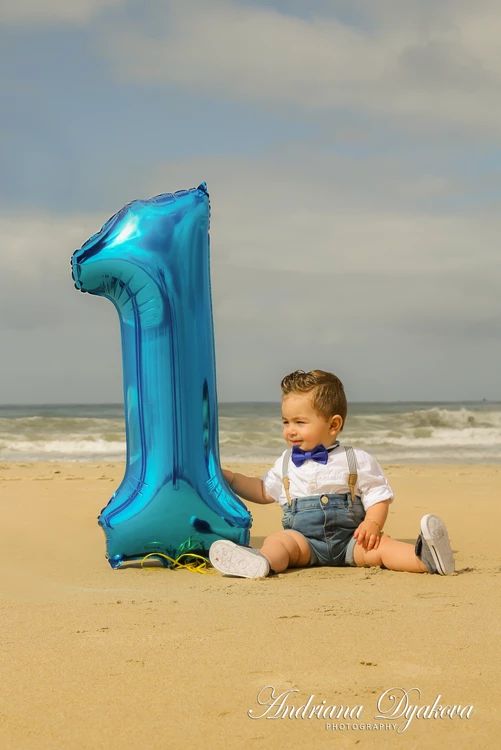 a little boy sitting on the beach with a blue number one balloon in front of him