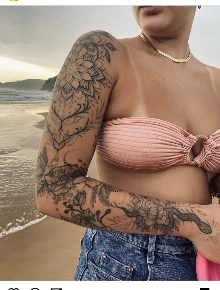 a woman with tattoos on her arm holding a baby in front of the ocean and beach