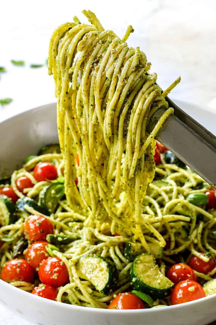 someone is using a fork to grab some noodles from a white bowl filled with tomatoes, zucchini and cucumbers