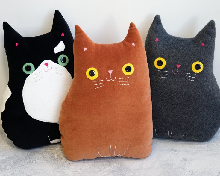 three pillows with cats on them are lined up against a white wall, one is black and the other has yellow eyes