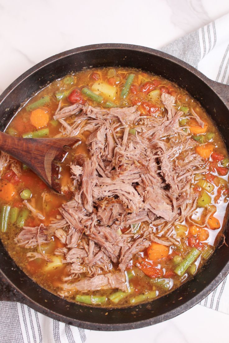 a skillet filled with meat and vegetables on top of a white table cloth next to a wooden spoon