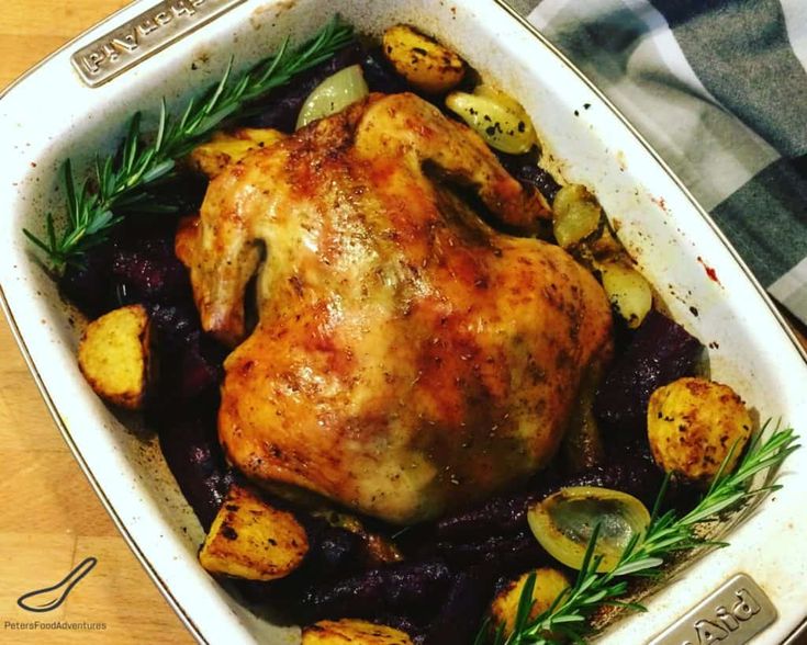 a roasted chicken in a casserole dish with potatoes and rosemary garnish