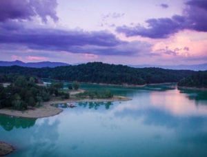 an aerial view of a lake surrounded by mountains and trees at dusk with clouds in the sky