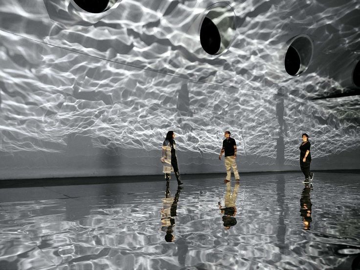 three people standing in front of a large white wall with circular lights hanging from it's ceiling