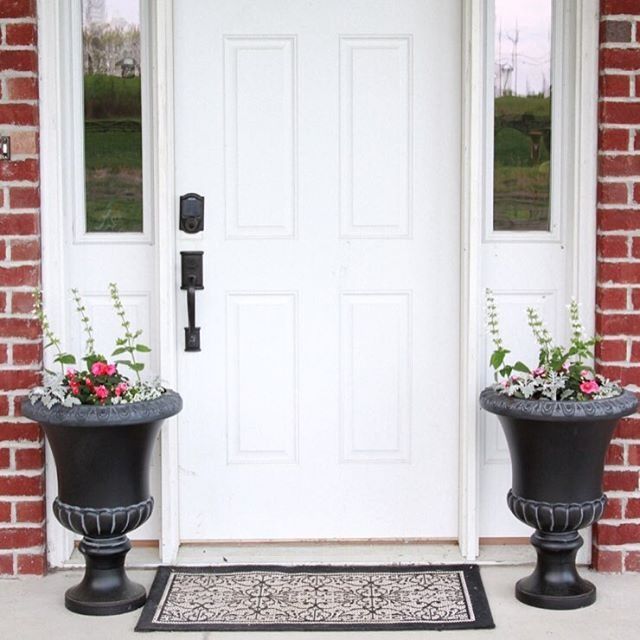 two black planters sitting in front of a white door with flowers on them and a welcome mat